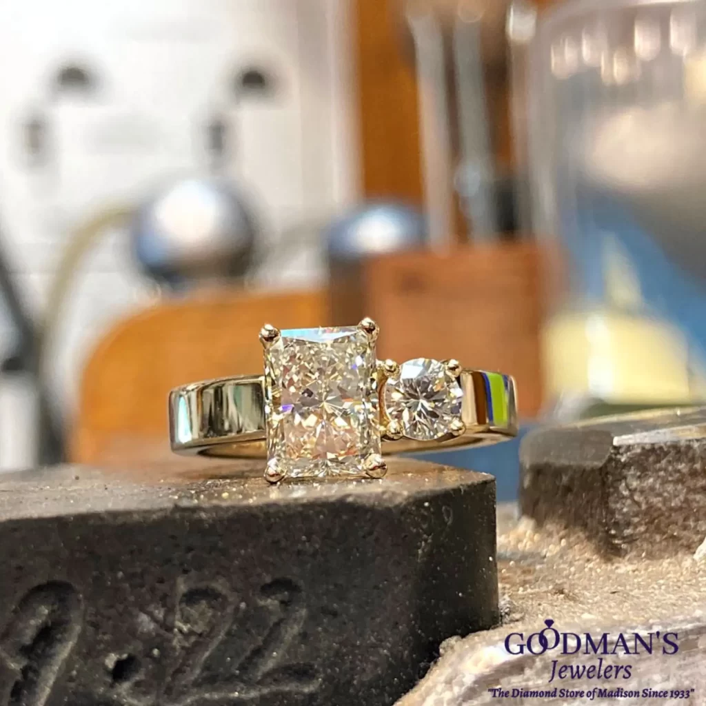A close-up of a two-stone diamond engagement ring, featuring a radiant-cut and round diamond, displayed in a jeweler’s workshop with tools in the background.